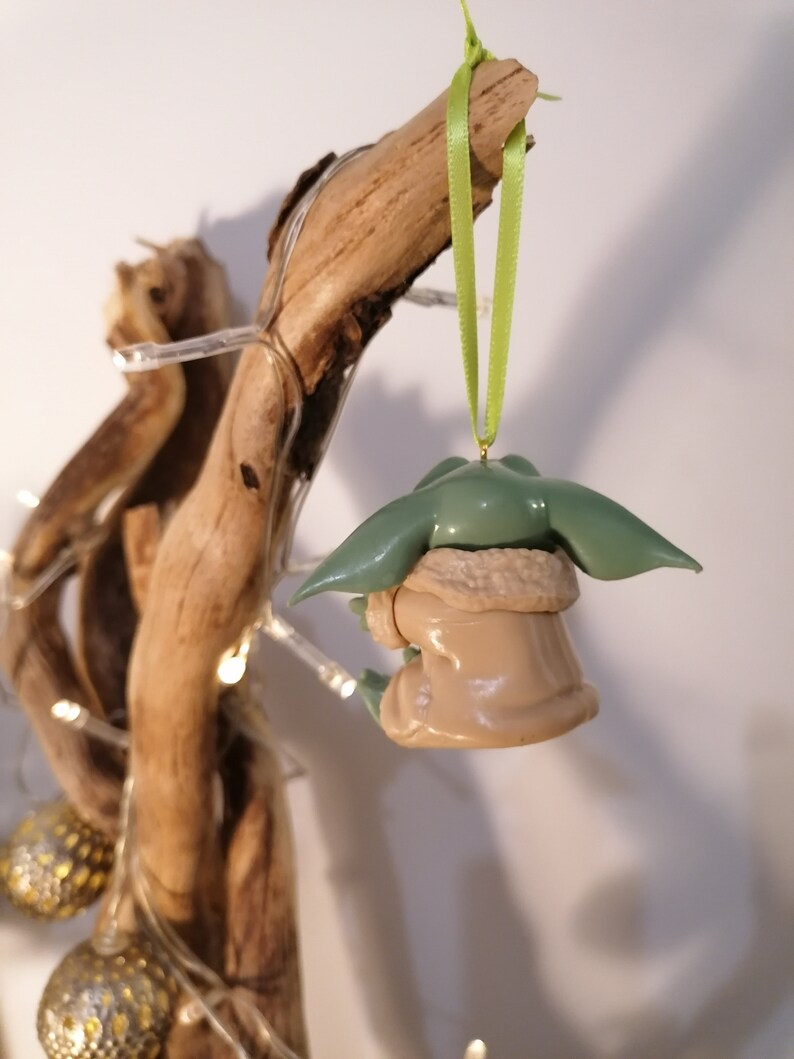 May include: A green and brown figurine of a character from the Star Wars franchise, hanging from a green ribbon on a branch of driftwood. The figurine is wearing a brown coat and has large ears.