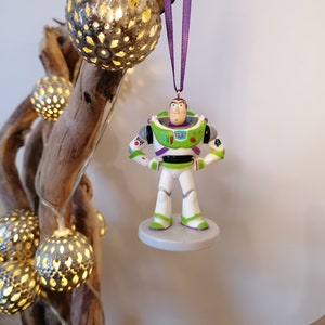 May include: A purple and white Christmas ornament featuring a plastic figurine of Buzz Lightyear from the Toy Story franchise. The ornament is hanging from a purple ribbon.