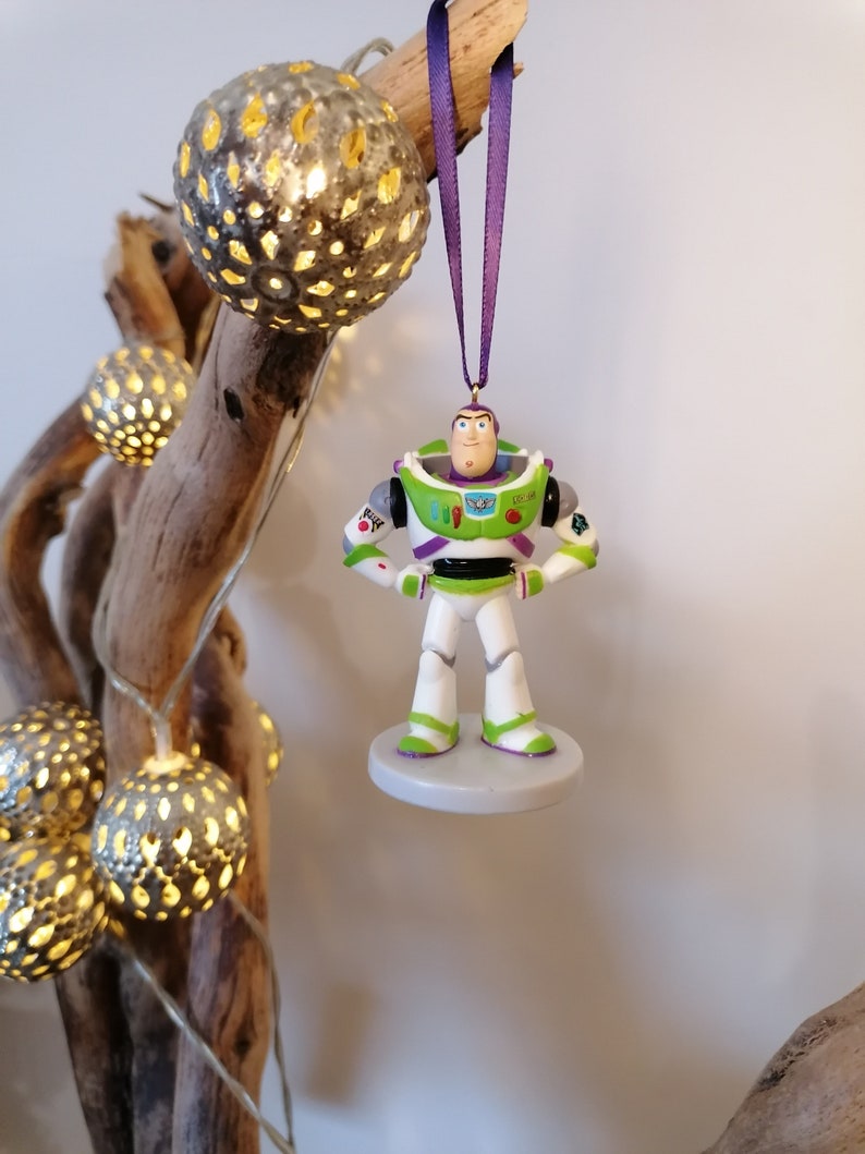 May include: A purple and white Christmas ornament featuring a plastic figurine of Buzz Lightyear from the Toy Story franchise. The ornament is hanging from a purple ribbon.