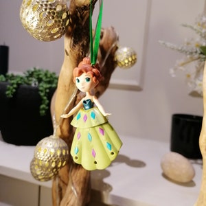 May include: A green and purple dress ornament featuring a cartoon character with brown hair and a blue bodice. The ornament is hanging from a green ribbon on a wooden branch.