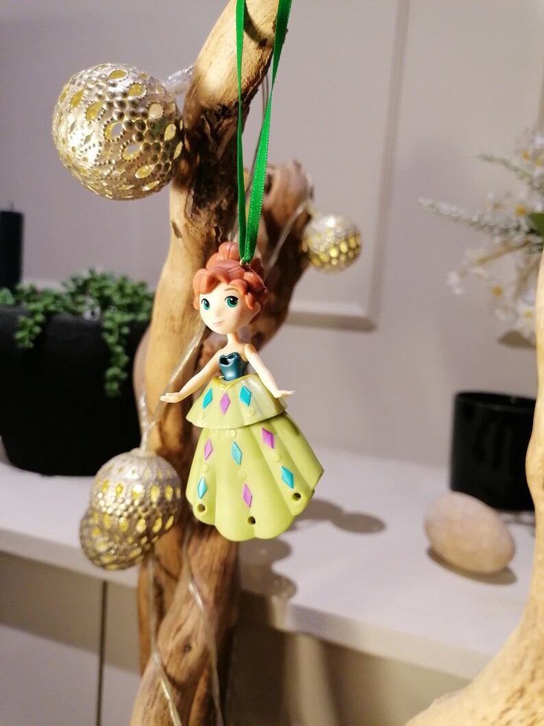 May include: A green and purple dress ornament featuring a cartoon character with brown hair and a blue bodice. The ornament is hanging from a green ribbon on a wooden branch.