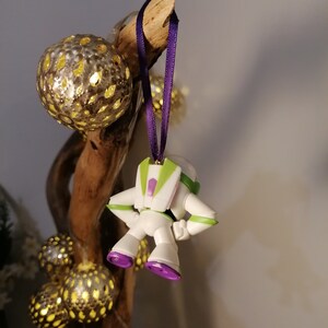 May include: A white and green plastic toy astronaut ornament with purple accents hanging from a branch with gold and white Christmas lights.