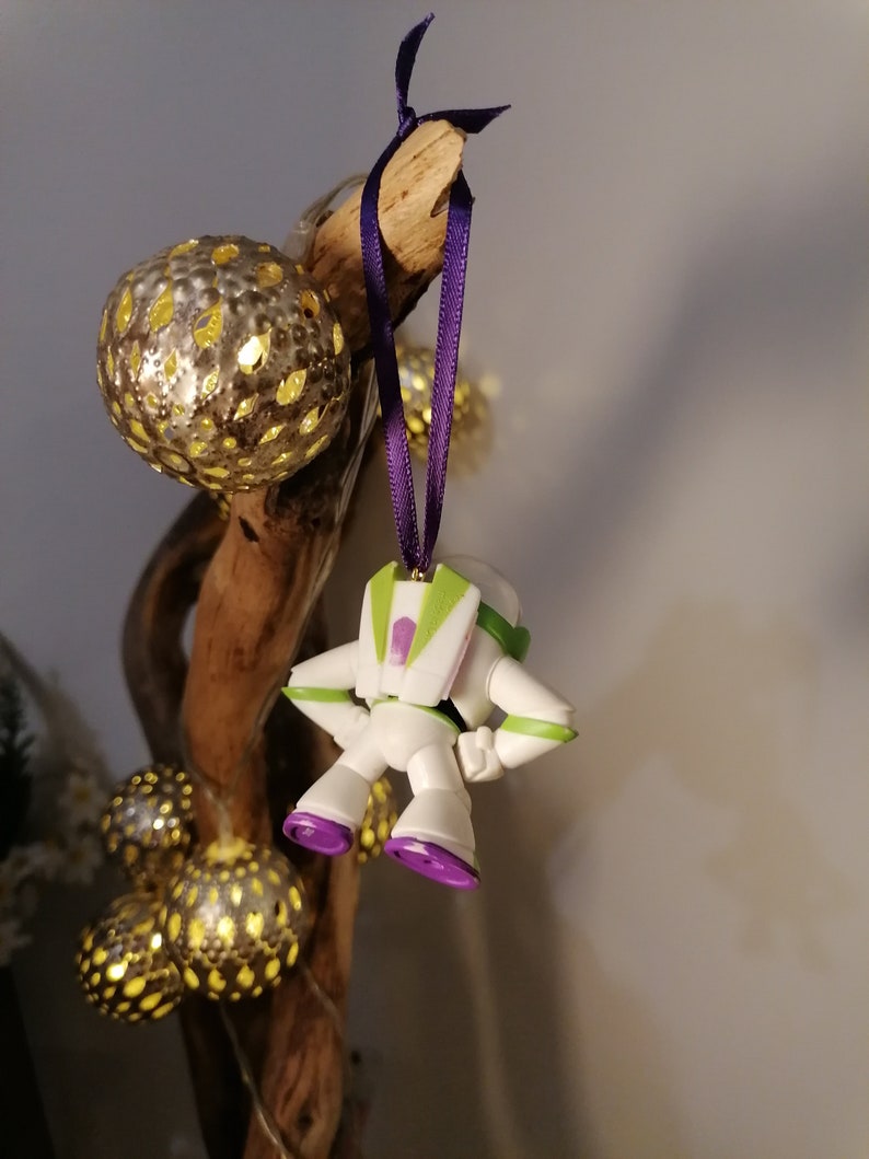 May include: A white and green plastic toy astronaut ornament with purple accents hanging from a branch with gold and white Christmas lights.