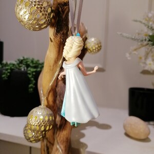 May include: A white and blue plastic figurine of a princess with blonde hair, wearing a white dress and a blue cape. The figurine is hanging from a branch with a silver ribbon.