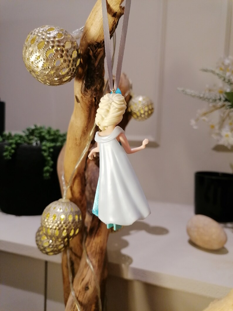 May include: A white and blue plastic figurine of a princess with blonde hair, wearing a white dress and a blue cape. The figurine is hanging from a branch with a silver ribbon.