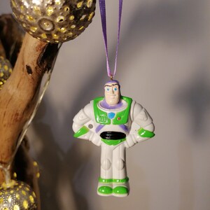Puede incluir: Un adorno de pl&aacute;stico blanco de Buzz Lightyear de la franquicia Toy Story. El adorno es verde, blanco y morado con un lazo dorado para colgar.