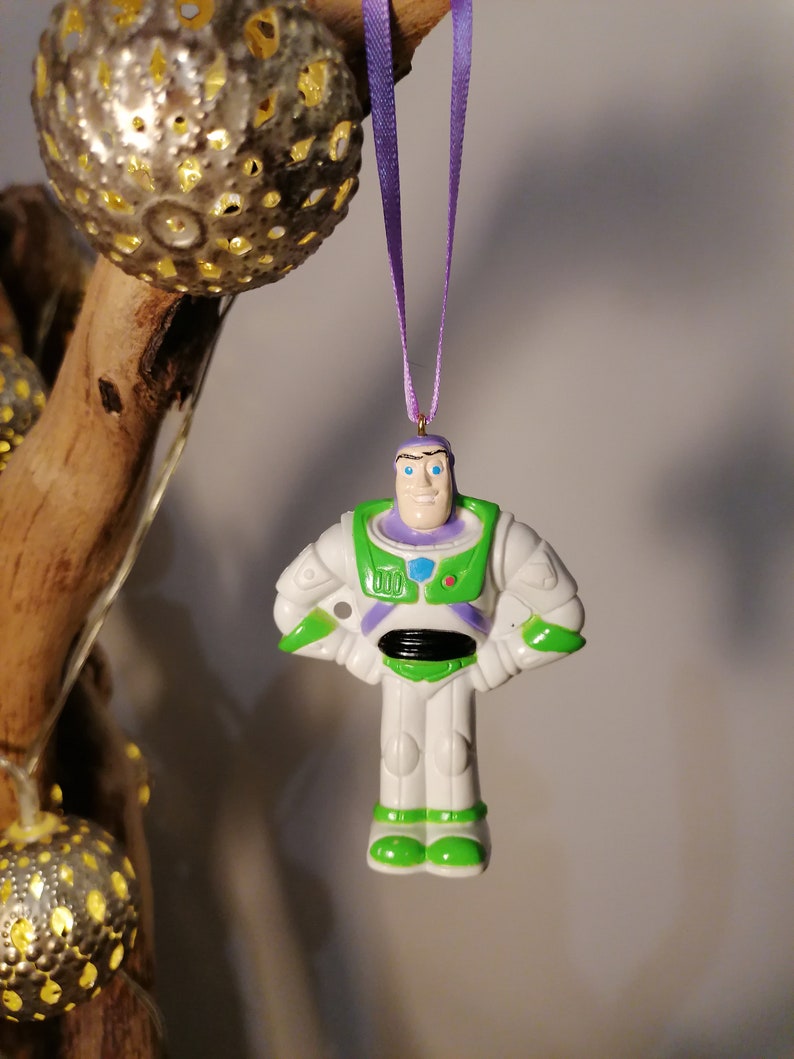 Puede incluir: Un adorno de pl&aacute;stico blanco de Buzz Lightyear de la franquicia Toy Story. El adorno es verde, blanco y morado con un lazo dorado para colgar.