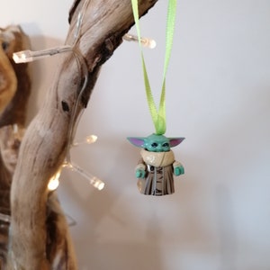 Puede incluir: Una figurita verde y marr&oacute;n de un personaje de la franquicia Star Wars, colgada de una cinta verde en una rama. La figurita es un adorno navide&ntilde;o.