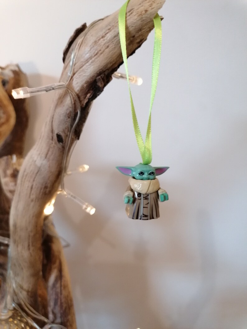 Puede incluir: Una figurita verde y marr&oacute;n de un personaje de la franquicia Star Wars, colgada de una cinta verde en una rama. La figurita es un adorno navide&ntilde;o.