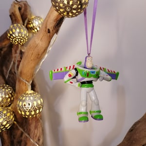 Peut inclure: Ornement de No&euml;l Buzz l'&Eacute;clair, v&ecirc;tu d'une combinaison spatiale blanche et verte avec des ailes violettes, suspendu par un ruban violet. L'ornement est accroch&eacute; &agrave; une branche d&eacute;corative, avec des orbes argent&eacute;s illumin&eacute;s et des guirlandes lumineuses.