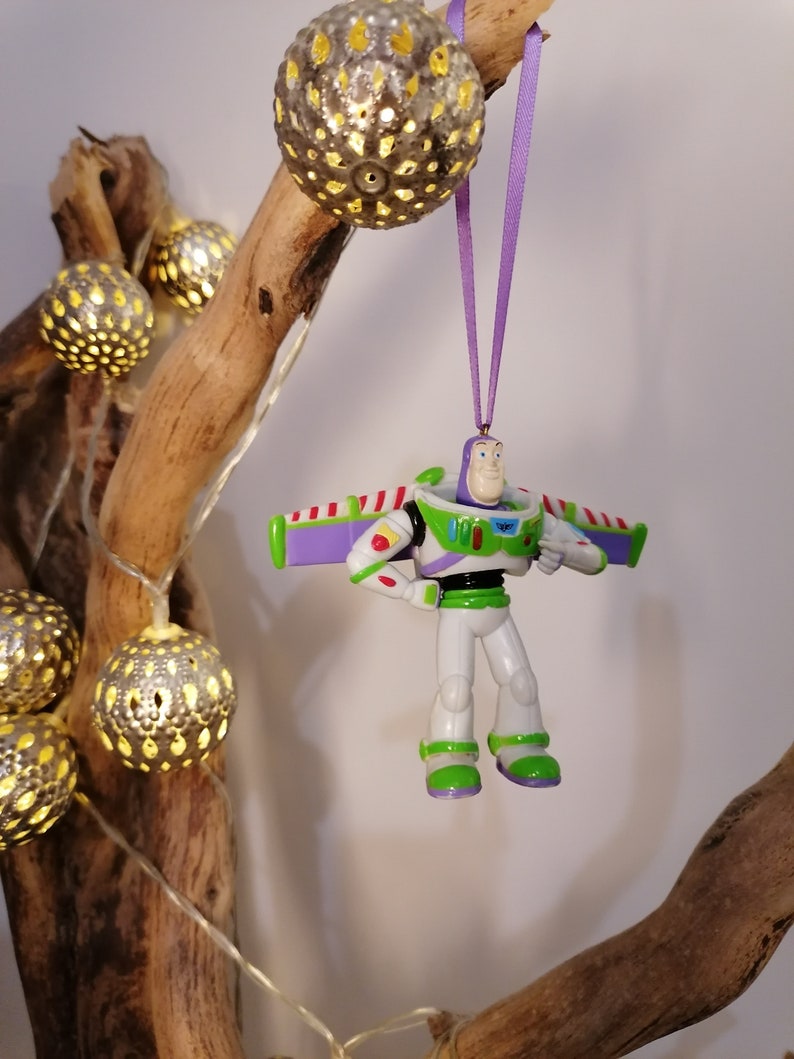 Peut inclure: Ornement de No&euml;l Buzz l'&Eacute;clair, v&ecirc;tu d'une combinaison spatiale blanche et verte avec des ailes violettes, suspendu par un ruban violet. L'ornement est accroch&eacute; &agrave; une branche d&eacute;corative, avec des orbes argent&eacute;s illumin&eacute;s et des guirlandes lumineuses.