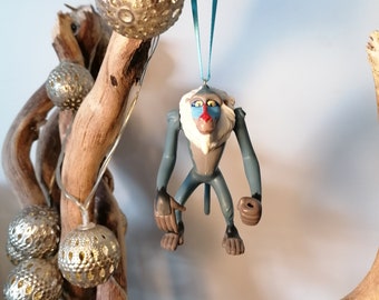 May include: A blue and gray monkey ornament with a red and blue face hanging from a blue ribbon. The ornament is hanging from a branch with silver ornaments.