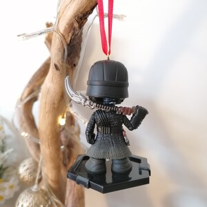 May include: Black and silver Star Wars ornament featuring Kylo Ren with a lightsaber. The ornament is hanging from a red ribbon on a wooden branch.