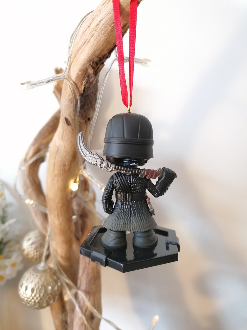 May include: Black and silver Star Wars ornament featuring Kylo Ren with a lightsaber. The ornament is hanging from a red ribbon on a wooden branch.