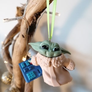 May include: A green and brown Christmas ornament featuring a character from the Star Wars franchise, The Mandalorian. The character is wearing a pink coat with a white fur collar and is holding a blue device with buttons.