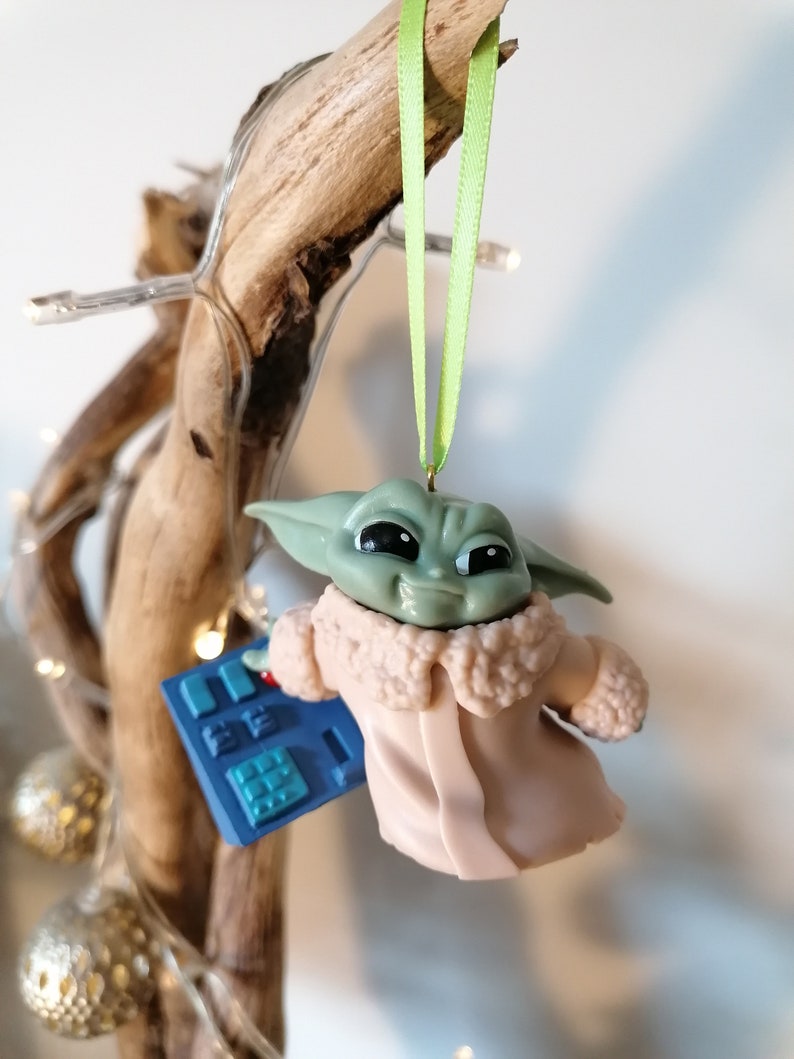 May include: A green and brown Christmas ornament featuring a character from the Star Wars franchise, The Mandalorian. The character is wearing a pink coat with a white fur collar and is holding a blue device with buttons.