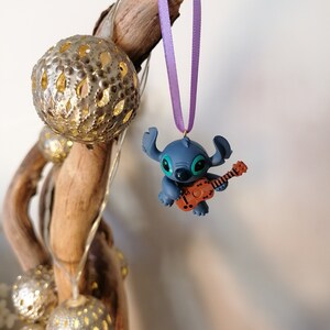 Op de afbeelding: Een blauwe Stitch-figuur ornament dat gitaar speelt, hangend aan een paars lint aan een houten tak.