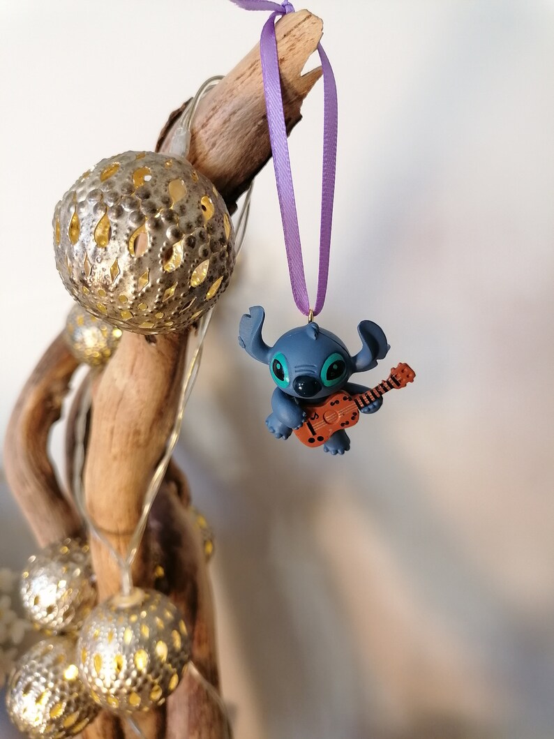Op de afbeelding: Een blauwe Stitch-figuur ornament dat gitaar speelt, hangend aan een paars lint aan een houten tak.