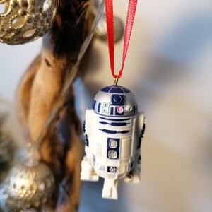 K&ouml;nnte beinhalten: Ein wei&szlig;er und blauer R2-D2 Star Wars Charakter Anh&auml;nger mit einem roten Band, das an einem Ast h&auml;ngt.
