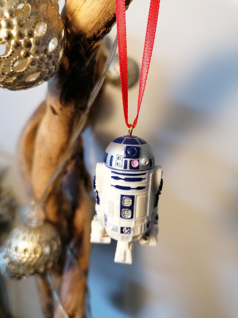 K&ouml;nnte beinhalten: Ein wei&szlig;er und blauer R2-D2 Star Wars Charakter Anh&auml;nger mit einem roten Band, das an einem Ast h&auml;ngt.