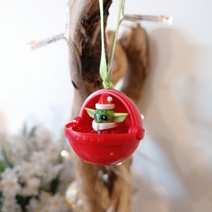 Pu&ograve; includere: Un ornamento natalizio rosso con un personaggio verde che indossa un cappello da Babbo Natale. Il personaggio &egrave; seduto in una piccola navicella spaziale rossa.