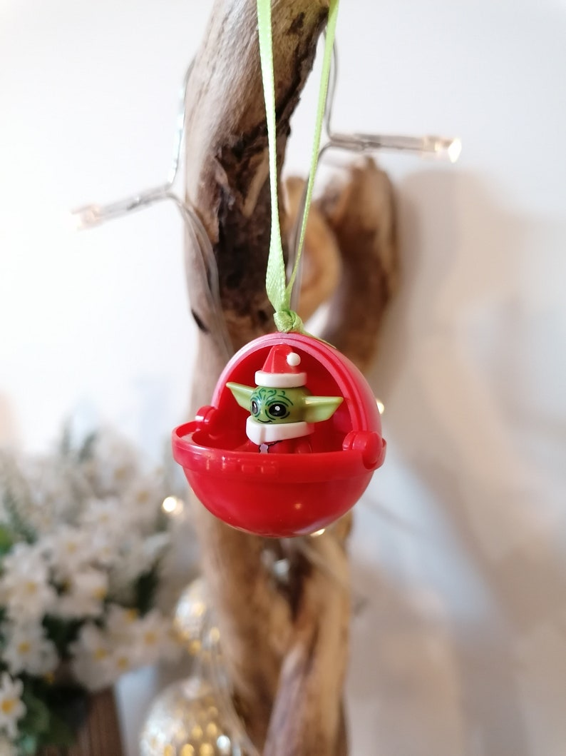Pu&ograve; includere: Un ornamento natalizio rosso con un personaggio verde che indossa un cappello da Babbo Natale. Il personaggio &egrave; seduto in una piccola navicella spaziale rossa.