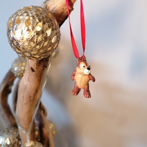 Pode incluir: Um ornamento de figurinha de esquilo marrom e bege com uma fita vermelha pendurada em um galho de madeira &agrave; deriva com ornamentos dourados.
