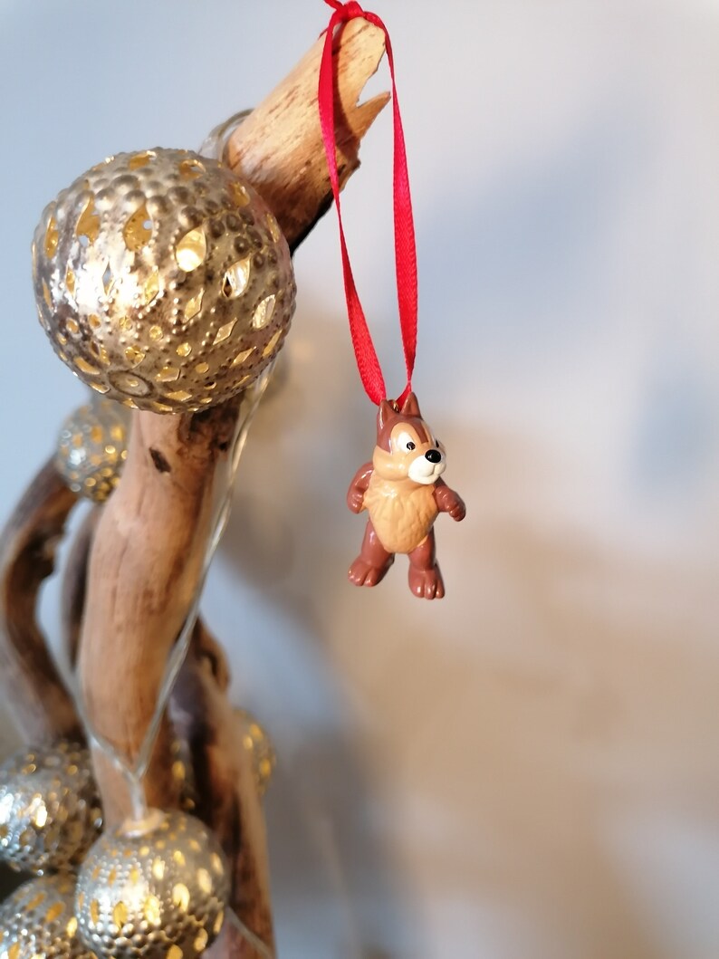 Pode incluir: Um ornamento de figurinha de esquilo marrom e bege com uma fita vermelha pendurada em um galho de madeira &agrave; deriva com ornamentos dourados.