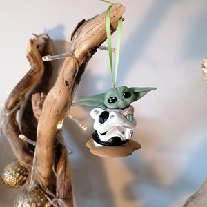 K&ouml;nnte beinhalten: Ein gr&uuml;ner und brauner Weihnachtsbaumschmuck mit einer Figur aus dem Star Wars-Franchise, The Mandalorian. Die Figur sitzt auf einem wei&szlig;en und schwarzen Objekt, m&ouml;glicherweise einem Helm.
