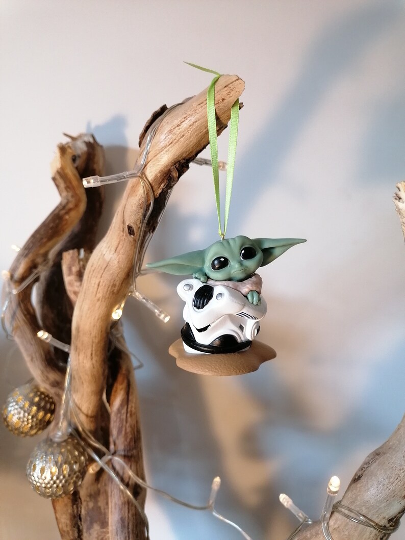 K&ouml;nnte beinhalten: Ein gr&uuml;ner und brauner Weihnachtsbaumschmuck mit einer Figur aus dem Star Wars-Franchise, The Mandalorian. Die Figur sitzt auf einem wei&szlig;en und schwarzen Objekt, m&ouml;glicherweise einem Helm.