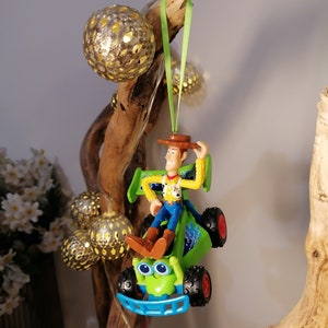 May include: A green toy car with a blue bumper and a figure of Woody from Toy Story driving it. The car is hanging from a branch with a green ribbon.