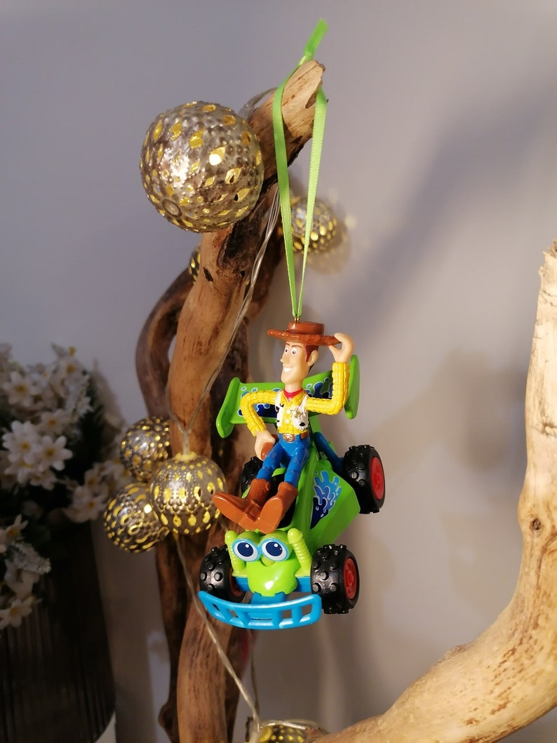May include: A green toy car with a blue bumper and a figure of Woody from Toy Story driving it. The car is hanging from a branch with a green ribbon.