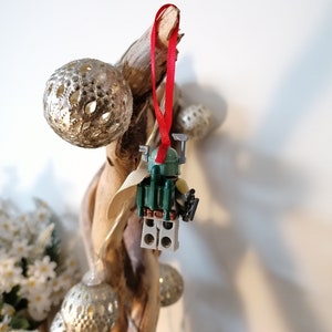 May include: A green and brown Lego Star Wars Boba Fett minifigure ornament hanging from a branch with a red ribbon. The ornament is decorated with silver and gold ornaments.