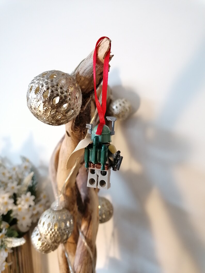 May include: A green and brown Lego Star Wars Boba Fett minifigure ornament hanging from a branch with a red ribbon. The ornament is decorated with silver and gold ornaments.