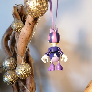 May include: Purple and pink character ornament with a flower on its head, hanging from a purple ribbon. The ornament is a cartoon character with a white tail and is wearing a purple dress.
