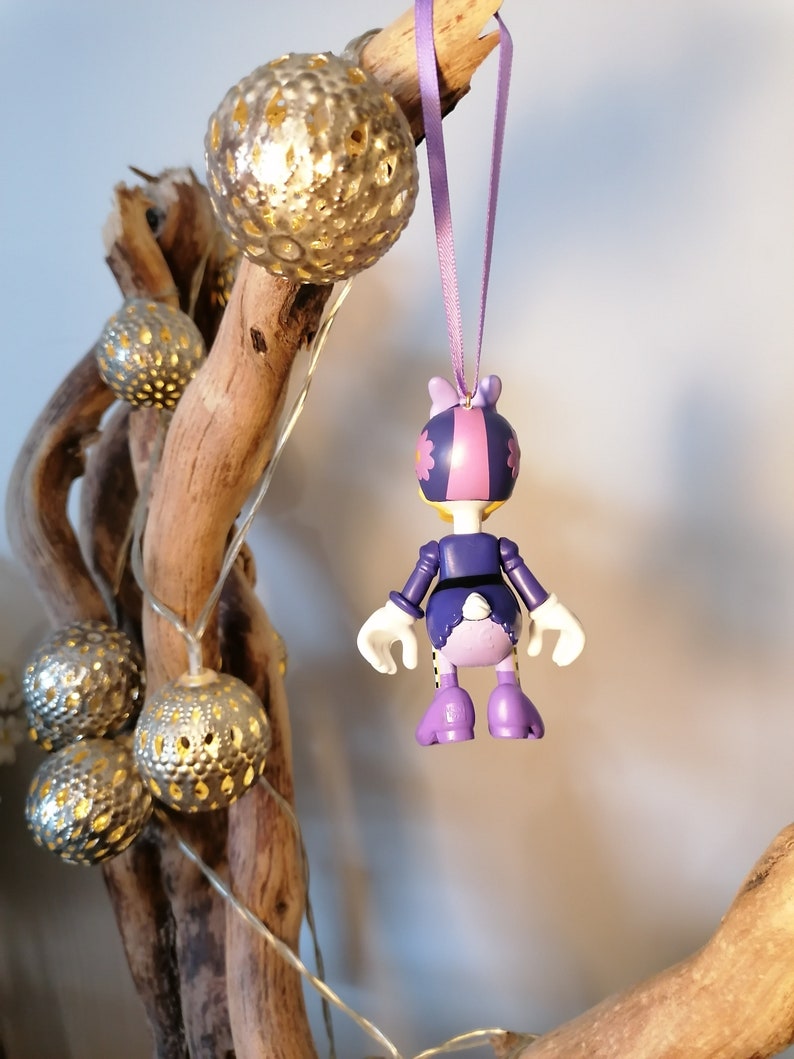 May include: Purple and pink character ornament with a flower on its head, hanging from a purple ribbon. The ornament is a cartoon character with a white tail and is wearing a purple dress.