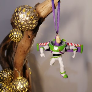 May include: A green and purple Buzz Lightyear Christmas ornament with wings, hanging from a purple ribbon on a wooden branch with decorative lights.