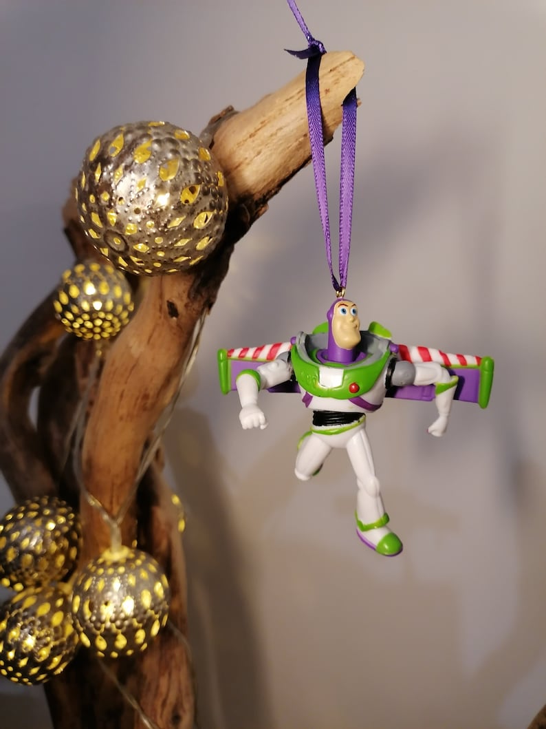 May include: A green and purple Buzz Lightyear Christmas ornament with wings, hanging from a purple ribbon on a wooden branch with decorative lights.