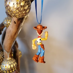 May include: A plastic ornament of Woody from Toy Story, wearing his signature cowboy hat and vest. He is holding his arm up in the air, as if he is waving. The ornament is hanging from a blue ribbon.