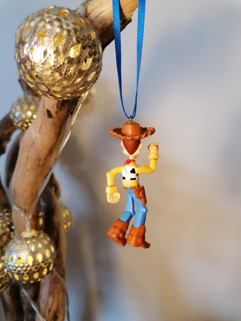 May include: A plastic ornament of Woody from Toy Story, wearing his signature cowboy hat and vest. He is holding his arm up in the air, as if he is waving. The ornament is hanging from a blue ribbon.