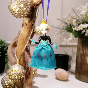 K&ouml;nnte beinhalten: Eine blaue und schwarze Plastikpuppe mit einer Krone, die ein blaues Kleid mit Schneeflockenmuster tr&auml;gt, das an einem violetten Band h&auml;ngt.