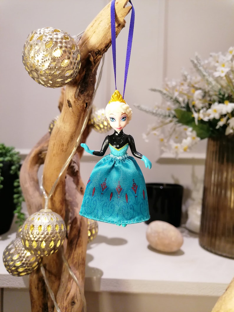 K&ouml;nnte beinhalten: Eine blaue und schwarze Plastikpuppe mit einer Krone, die ein blaues Kleid mit Schneeflockenmuster tr&auml;gt, das an einem violetten Band h&auml;ngt.
