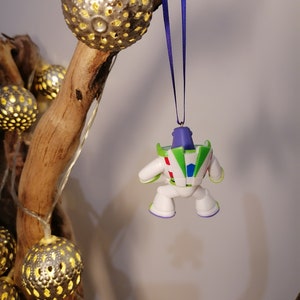 May include: A white and green Buzz Lightyear Christmas ornament with a purple ribbon hanging from a branch with gold string lights.