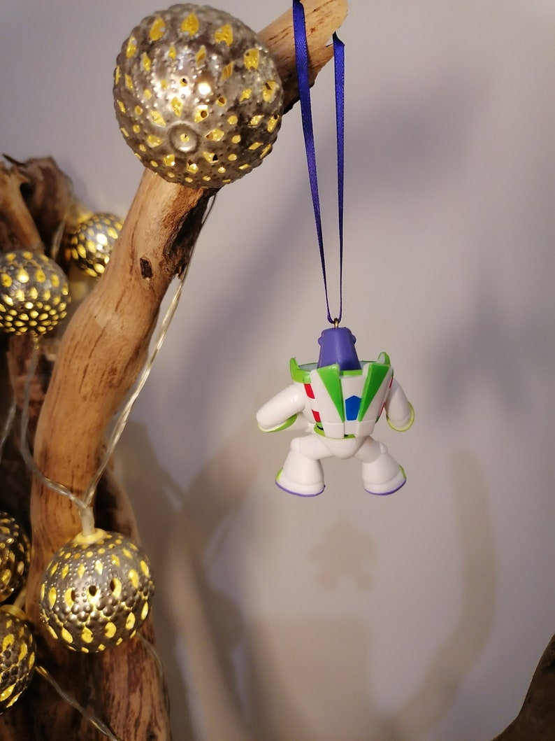 May include: A white and green Buzz Lightyear Christmas ornament with a purple ribbon hanging from a branch with gold string lights.
