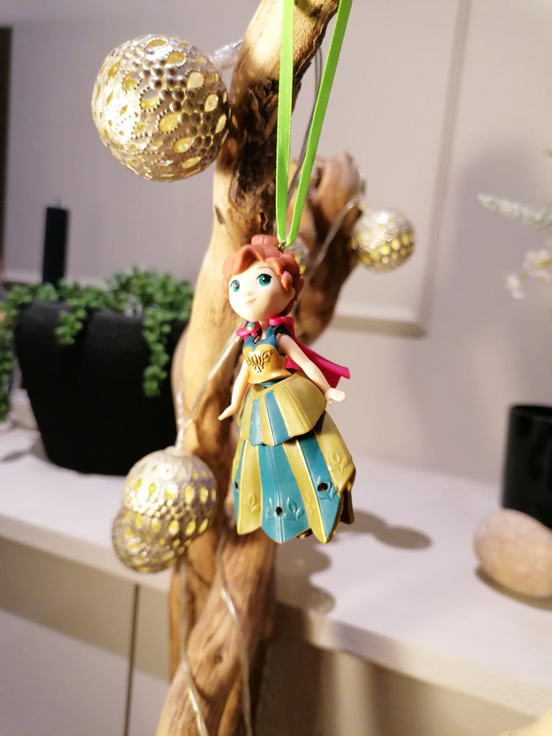 Pu&ograve; includere: Una piccola figurina di plastica di una ragazza che indossa un vestito blu e oro con un mantello rosso appeso a un nastro verde su un ramo con luci.