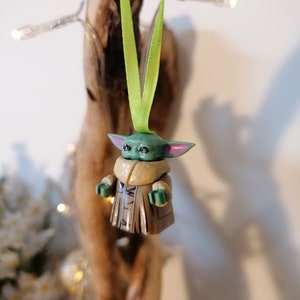 Puede incluir: Una figurita verde y marr&oacute;n de un personaje de la franquicia Star Wars, colgada de una cinta verde. La figurita lleva un abrigo marr&oacute;n y tiene orejas grandes.