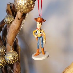 Op de afbeelding: Een rode draad hangt aan een houten tak met een witte ronde basis en een beeldje van Woody uit Toy Story. Woody draagt zijn kenmerkende bruine hoed, zijn gele shirt, zijn blauwe spijkerbroek en bruine laarzen.