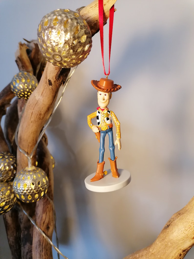 Op de afbeelding: Een rode draad hangt aan een houten tak met een witte ronde basis en een beeldje van Woody uit Toy Story. Woody draagt zijn kenmerkende bruine hoed, zijn gele shirt, zijn blauwe spijkerbroek en bruine laarzen.