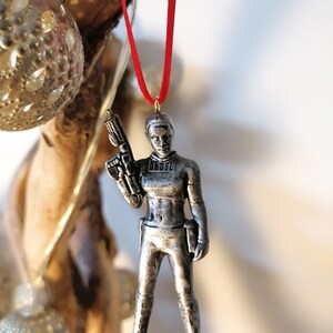May include: A silver-coloured ornament of a female figure holding a gun. The ornament is hanging from a red ribbon.