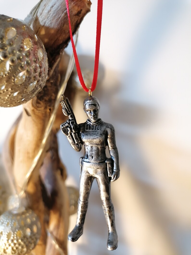 May include: A silver-coloured ornament of a female figure holding a gun. The ornament is hanging from a red ribbon.